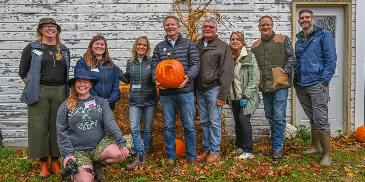 Group of people standing in front of a weathered white wooden building with a sign that reads ‘North Farm.’ They are posing outdoors on grass with fallen leaves, and one person is holding a large orange pumpkin. Hay bales, cornstalks, and additional pumpkins are arranged as decorations near the building