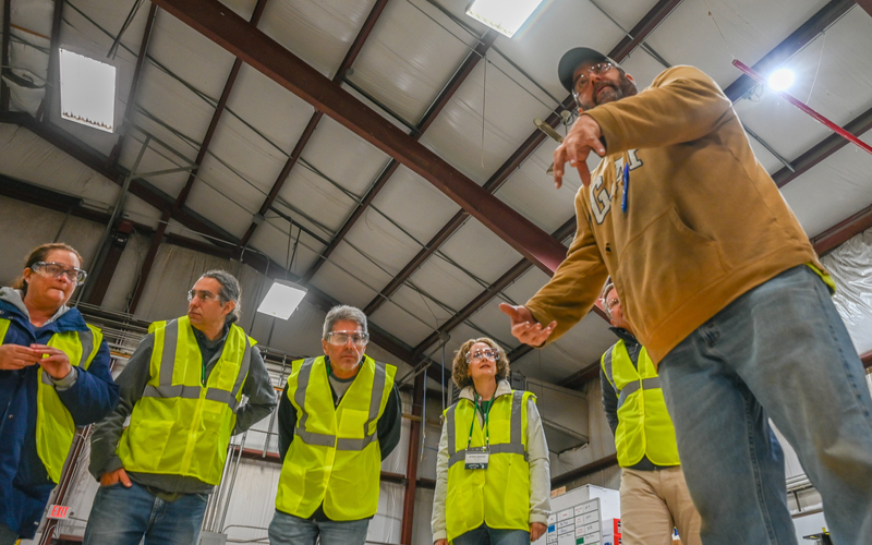A group of people wearing bright yellow safety vests stand inside a large industrial building with a high metal ceiling and exposed beams. One person in the foreground, dressed in a tan hoodie and a black cap, gestures with one hand while speaking to the group. The background includes structural elements, fluorescent lights, and a whiteboard with notes, suggesting a tour or safety briefing in a warehouse or manufacturing facility.