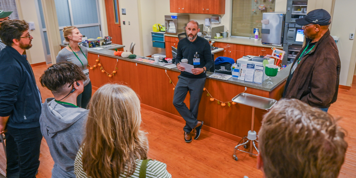 A group of people standing in a hospital or clinic break room, listening to a person who is seated on the counter and holding papers. The room has wooden cabinets, medical supplies on the counter, and orange garland decorations. There is a metal rolling tray and various equipment visible in the background