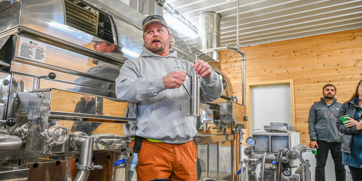 A person wearing an ash-gray hoodie and orange pants holds a metal container inside a facility with stainless steel equipment and wooden walls. The setting appears to be a production or processing area, with large machinery, pipes, and a countertop in the foreground. Other individuals stand nearby observing, and shelves with various items are visible in the background.