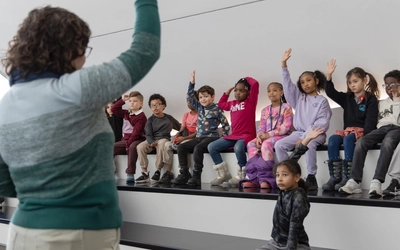 A teacher stands in front of a group of young students sitting on tiered benches, engaging them in a lesson.