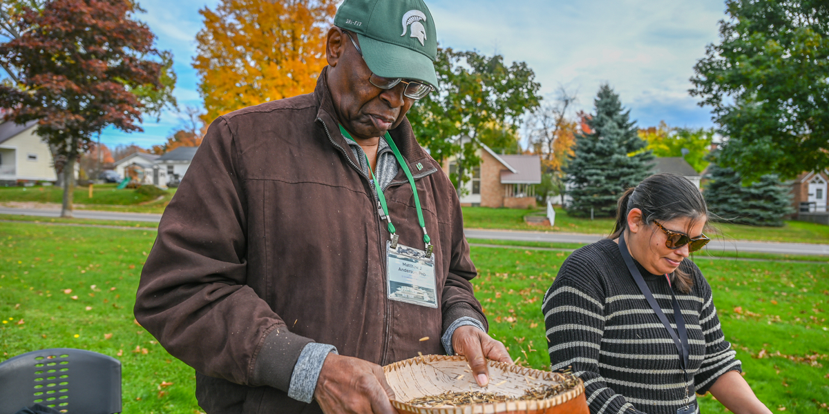 Two people stand outdoors on a grassy area with autumn trees and houses in the background. Both are holding wooden trays filled with seeds or grains, appearing to demonstrate or sort them. One person wears a dark jacket and green cap with a Spartan logo, while the other wears a striped sweater. Nearby is a black chair and a bag on the ground.