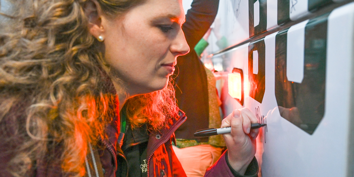 A person writes on the side of a large white vehicle using a black marker. The vehicle has bold black lettering, and the writing appears to be a signature or message added to the surface. The scene is illuminated by a red light reflecting on the person’s hair and jacket, suggesting nearby vehicle lights or brake lights. The background includes part of another arm raised toward the vehicle.