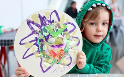 A young child wearing a green dinosaur hoodie sits at a table holding up a round art project decorated with colorful tissue paper, foil, and purple crayon scribbles.