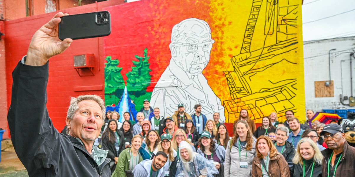A large group of people pose for a photo in front of a colorful mural painted on a brick wall. The mural features bright red, orange, and yellow sections with an outlined figure and architectural elements, along with green trees and a blue waterfall on the left side. One person in the foreground holds up a smartphone to take a selfie with the group