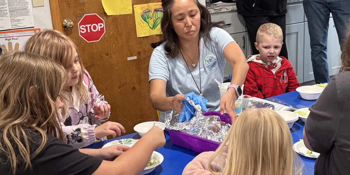 Several children are seated around a blue table in a classroom, eating from white bowls filled with salad. An adult at the center of the table is serving food from a foil-covered container placed in a purple tray. The background shows a wooden door decorated with colorful paper hearts and drawings, a small red “STOP” sign, and shelves with stacked papers. Additional adults are standing near the counter behind the group.