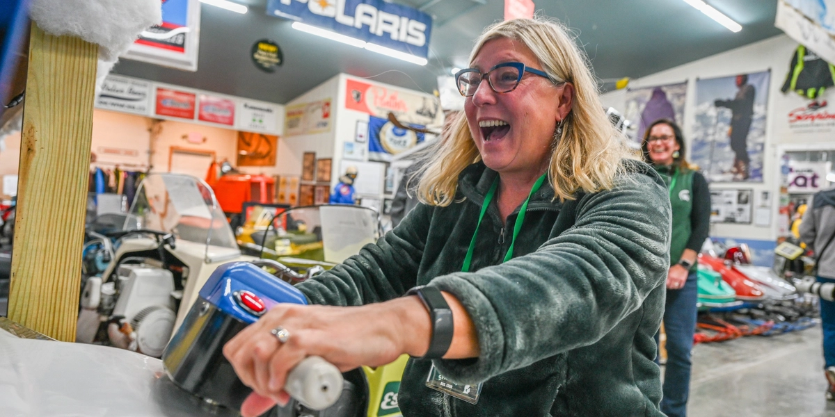 A person sits on a vintage snowmobile inside a museum filled with snowmobiles and related memorabilia. The individual grips the handlebars of a snowmobile arcade game, which has a white front panel and a blue control area. The background features multiple colorful snowmobiles, posters, and banners, including a prominent “Polaris” sign hanging from the ceiling. The setting is well-lit with fluorescent lights, and the floor is concrete.