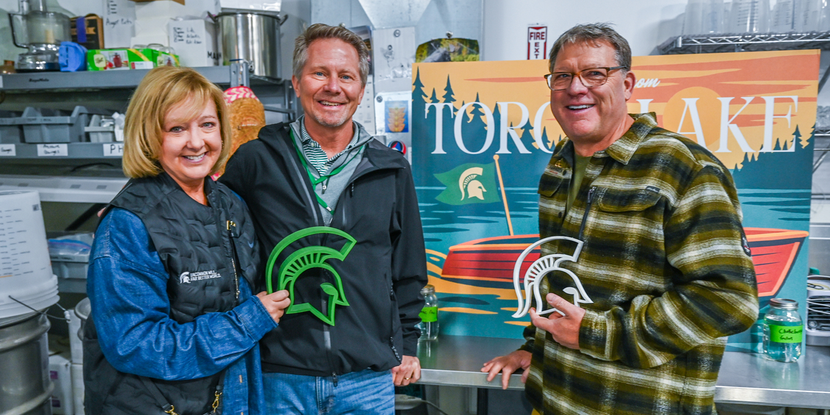 Three people stand in a commercial kitchen area, posing for a photo while holding green and white Spartan helmet cutouts. Behind them is a colorful sign that reads “Torch Lake” with an illustration of a red boat on water and trees in the background. The countertop in front of them has bottles and cans, and shelves with kitchen supplies are visible in the background.