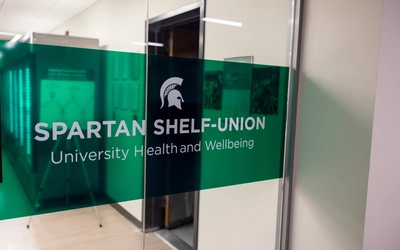 Photo of a window with a green clear semi transparent banner with white lettering saying "Spartan Shelf- Union, University health and wellbeing"