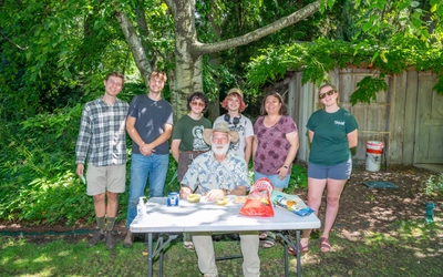 Six people gathered outdoors around a table with snacks and drinks. Five of them stand around one who is sitting. Trees, bushes, and a wooden fence are in the background. Faces are blurred for privacy.