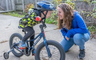 Gabrielle Ekstrum kneels beside son Mattias on training bike in driveway, both smiling.