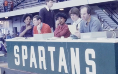 A group of people sit behind a long table at an indoor sports venue. The table has a large green front panel with the word “SPARTANS” printed in bold white letters
