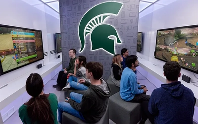 A modern gaming lounge where a group of people sit on stools playing video games on large wall‑mounted screens, with a prominent Spartan helmet logo on the back wall.