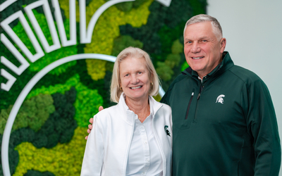 Two people stand side by side in front of a green and black moss wall featuring a large white Michigan State University Spartan helmet design. One person is wearing a white jacket with an MSU logo, and the other is wearing a dark green quarter‑zip pullover with an MSU logo. The person on the right has their arm around the person on the left. The setting appears bright and modern