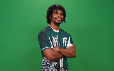 A varsity esports player wearing a green t-shirt jersey with a Spartan logo crosses his arms and smiles for the camera in front of a green screen