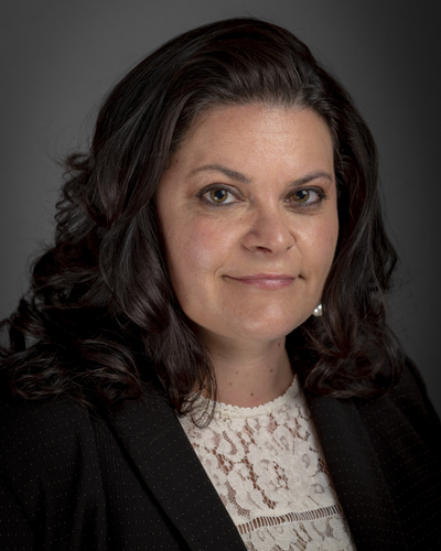 Person in a dark blazer and a white lace top posing for a portrait against a dark background, with shoulder‑length wavy hair.