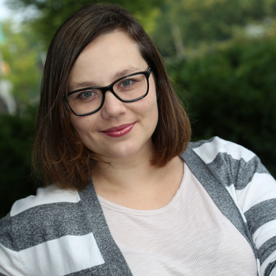 Person outdoors wearing a light-colored top and a gray-and-white striped cardigan, standing in front of greenery.