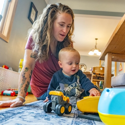 Gabrielle Ekstrum and baby Lukas play on rug with toys beneath a dining table at home.