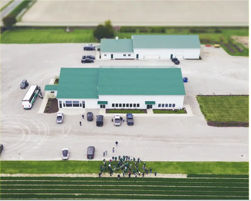 Aerial view of a research facility with green roofs, parked vehicles and a gathered crowd in a farm field.