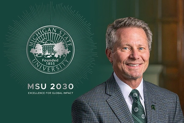 Portrait of a President Guskiewicz smiling in front of a green background beside the Michigan State University seal with "MSU 2030 EXCELLENCE FOR GLOBAL IMPACT" written below