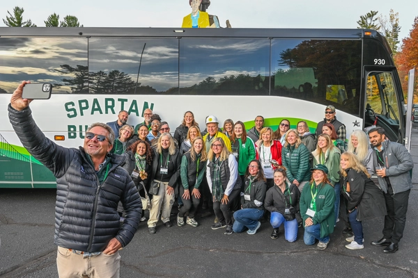 A large group of people pose for a photo in front of a white and green tour bus labeled “Spartan Bus Tour.” One person in the foreground holds up a smartphone to take a selfie with the group. The bus is parked on an asphalt lot surrounded by trees with autumn foliage, and a colorful wood figure is visible behind of the bus.