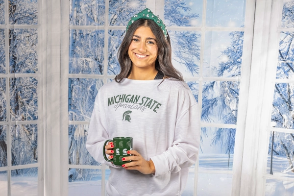 Woman in MSU sweatshirt holding Spartan mug in winter-themed studio setting.