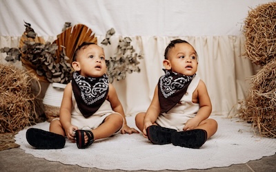 Young infant twins pose in front of autumnal decore with handkerchiefs and white outfits with black sandals