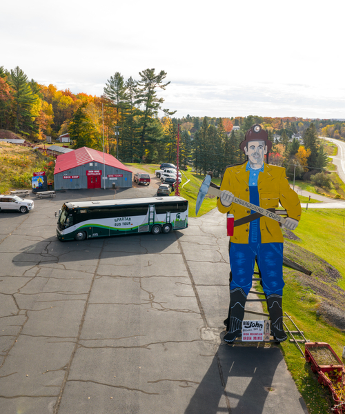 A large roadside wooden figure of a miner holding an axe stands on a paved area near a red-roofed building and a parked tour bus. The background features a winding road, trees with autumn foliage, and a bright sky.