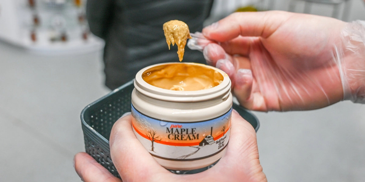 A close-up of a gloved hand holding a small plastic spoon with a dollop of thick maple cream above an open jar labeled “Pure Maple Cream.” The jar features an illustration of a snowy landscape with trees and a sugar shack. The background shows a gray countertop and blurred shelves with various items.