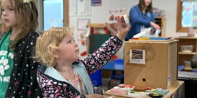 Two children are standing at a classroom activity station with wooden shelves. One child is holding up a puzzle piece while looking at a table covered with colorful wooden puzzles, including shapes and a picture of a basket of fruit. The other child stands nearby wearing a green shirt with a paw print design. In the background, there are educational posters on the wall and a person near a window organizing materials.