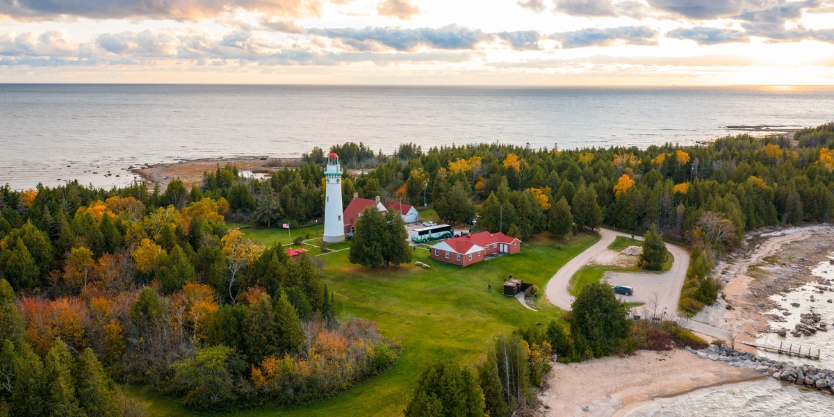 An aerial view of a scenic lakeshore featuring a historic lighthouse surrounded by dense green and autumn-colored trees. Several red-roofed buildings sit near the lighthouse on a grassy clearing, with a winding road leading to a small parking area. The shoreline includes sandy and rocky sections, and the expansive lake stretches to the horizon under a partly cloudy sky with soft sunlight