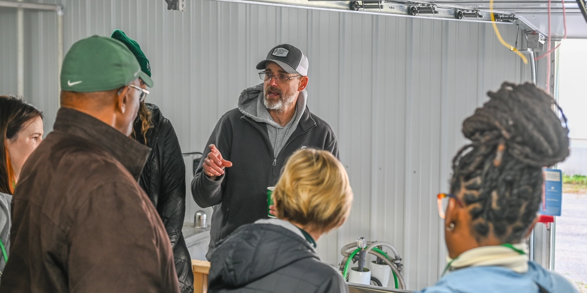 A group of people gathered inside a metal-walled facility, listening to a person in a dark jacket and baseball cap who appears to be explaining something. The group is standing around a workstation with pipes and hoses visible in the background, suggesting an industrial or agricultural setting.