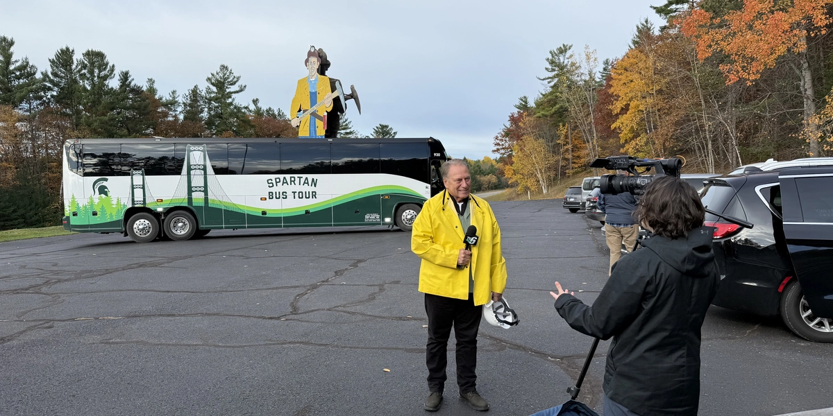 Tom Izzo wears a bright yellow jacket and stands in a parking lot holding a camera, while another person with a video camera films nearby. Behind them is a large tour bus labeled “Spartan Bus Tour” with a green and white design, and a tall statue of a lumberjack holding an axe is visible in the background. Several cars are parked along the edge of the lot, and colorful autumn trees line the scene under a cloudy sky.