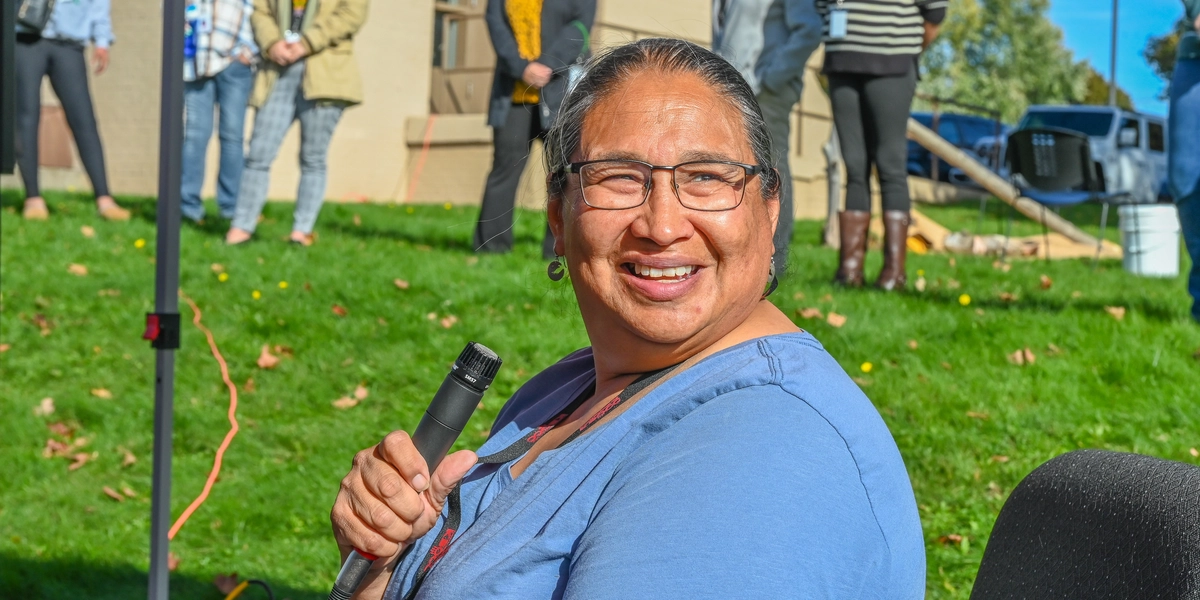 A person seated outdoors holding a microphone, with a group of people standing on a grassy area in the background near a beige building. The scene appears to be part of an event or gathering, with clear skies and sunlight casting shadows on the ground.