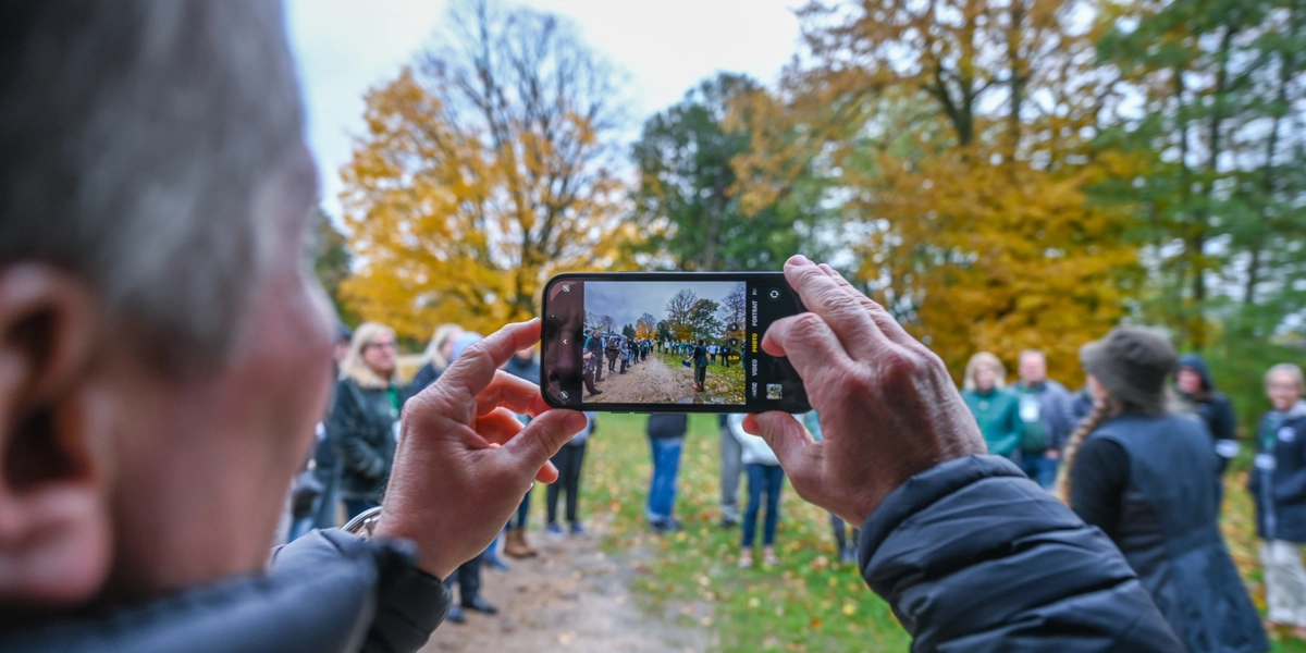A person holds a smartphone horizontally to take a photo of a group of people standing outdoors on a dirt path surrounded by autumn trees with colorful foliage. The phone screen shows the group clearly, while the background includes scattered leaves and tall trees in shades of green, yellow, and orange.