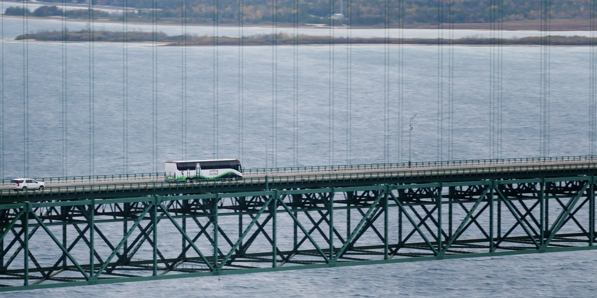 A green and white tour bus labeled “Spartan Bus Tour” drives across the Mackinac Bridge. A white car is traveling ahead of the bus. In the background, there is a shoreline with trees showing autumn colors and a cloudy sky above.