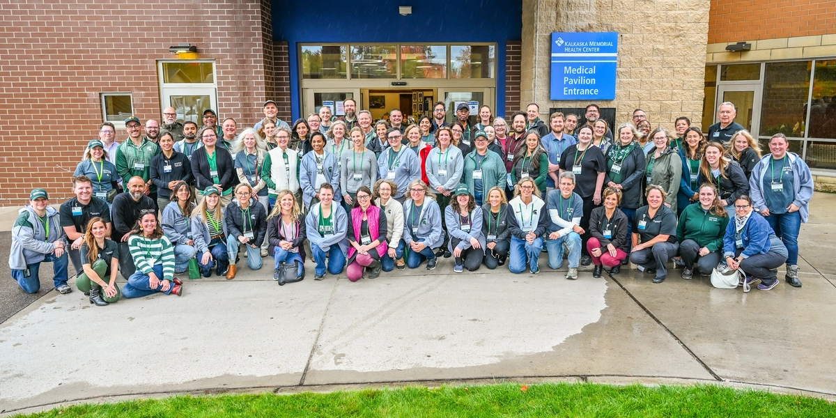 A large group of people posing for a photo outside a building entrance with a blue sign that reads “Medical Pavilion Entrance.” The group is standing and kneeling on a sidewalk in front of the entrance, wearing casual outdoor clothing, many in green and gray tones. The background shows brick and stone walls, glass doors, and windows.
