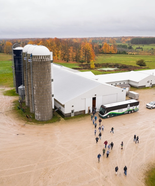 Aerial view of a large white farm building with two silos and a green-and-white tour bus parked outside. A group of people walk across a muddy lot toward the building, surrounded by green fields and autumn-colored trees under a cloudy sky.
