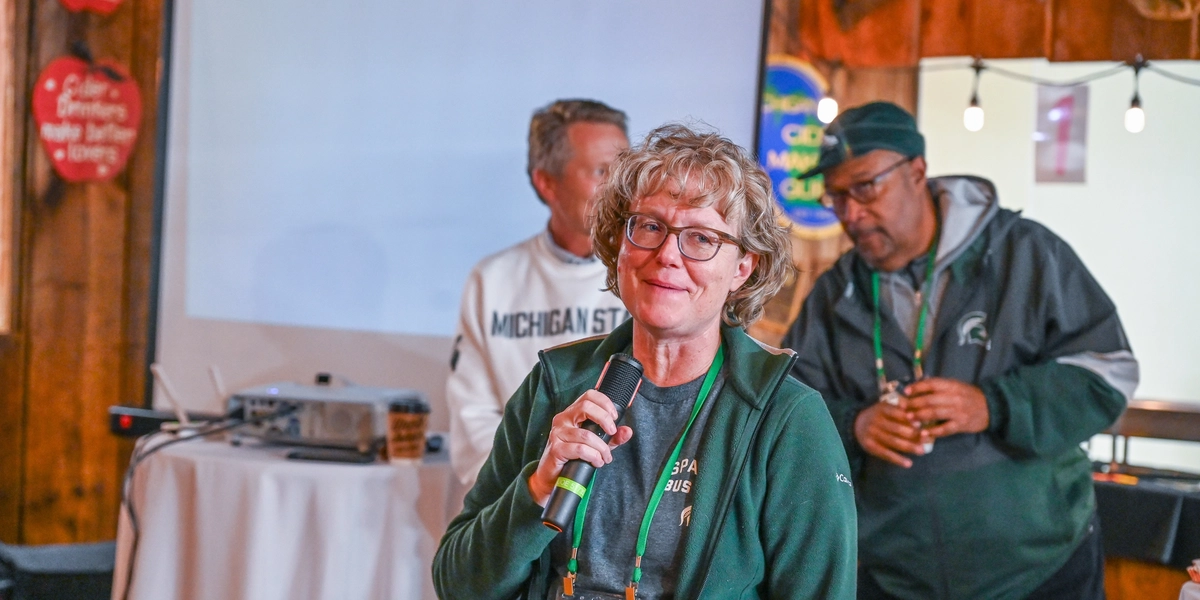 A person speaks into a microphone while standing indoors in front of a projector screen. The individual is wearing a green jacket with a lanyard and name badge. Behind the speaker, two other people are visible, one wearing a Michigan State sweatshirt and another in a dark jacket. The setting includes wooden walls, string lights, and heart-shaped wall decorations, creating a rustic atmosphere. A small round table with a projector and cup is positioned near the screen.