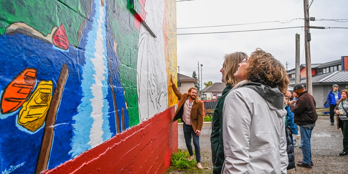 A group of people stand outdoors near a large, colorful mural painted on the side of a building. The mural features bright designs including a waterfall and kayaks in vivid colors. One person is closely observing the artwork, while others gather nearby in a gravel area with utility poles and buildings in the background.