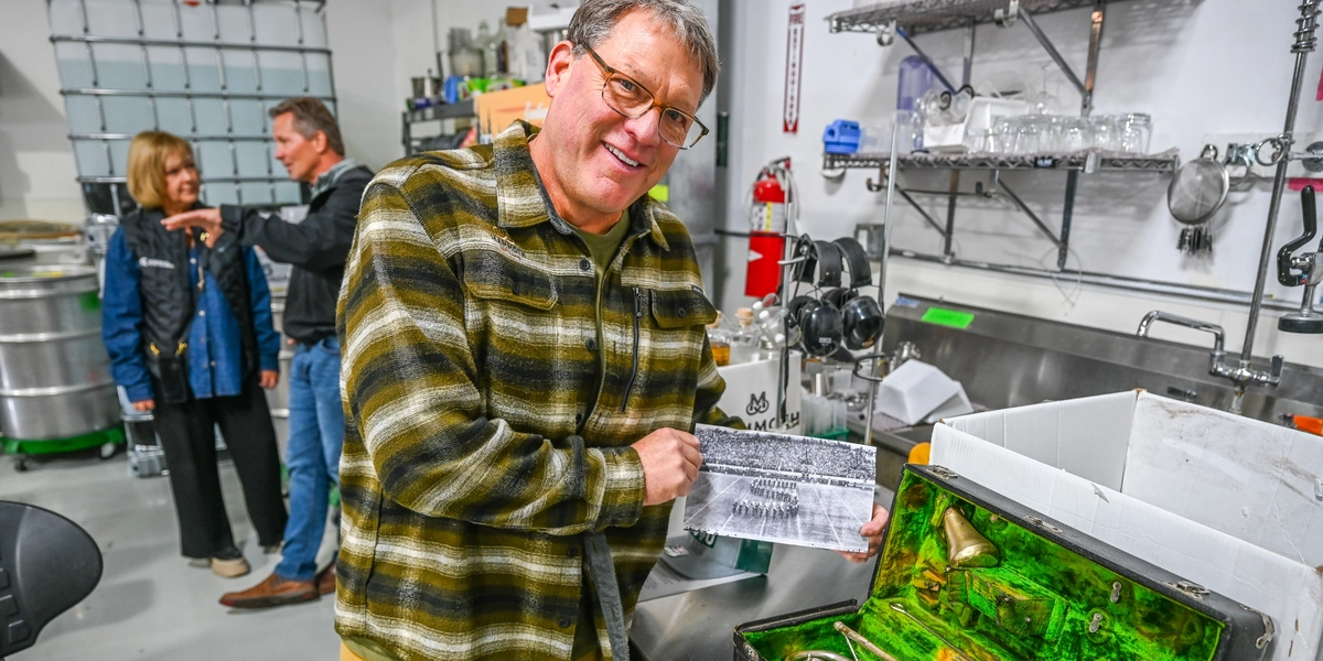 A person wearing a plaid shirt and yellow pants stands in a stainless-steel kitchen area, holding a black-and-white photo in one hand and pointing toward an open case containing an old trumpet. In the background, shelves with glassware, kitchen supplies, and a fire extinguisher are visible, along with two other people near large metal containers.