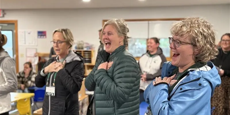 A group of adults standing inside a brightly lit classroom with wooden floors and colorful chairs. They are wearing jackets and name badges, and each person has both hands placed over their chest. Two of them have big smiles. In the background, there are shelves with educational materials and a large window letting in natural light.
