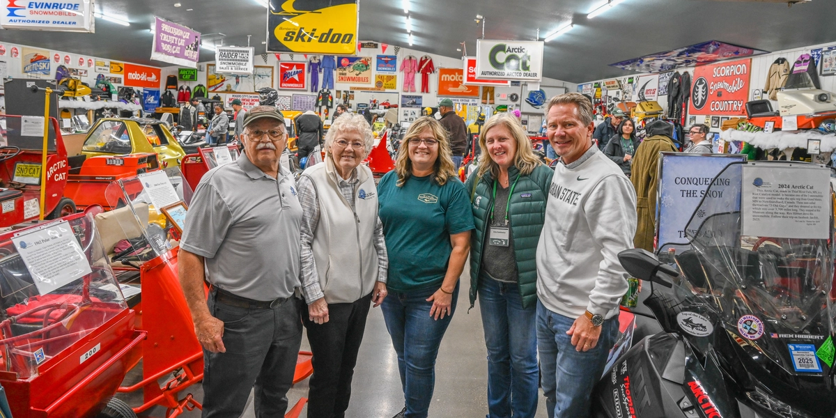 Five people stand together inside a snowmobile museum filled with vintage snowmobiles and memorabilia. Brightly colored snowmobiles, including red and black models, are displayed around them. The background features numerous signs and banners for brands such as “John Deere Snowmobiles,” “Ski-Doo,” and “Evinrude,” along with shelves of merchandise, posters, and informational displays. The setting is well-lit with overhead fluorescent lights.