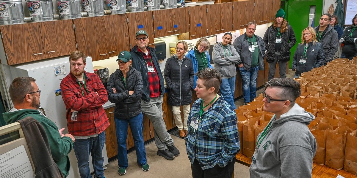 A group of people standing inside a room with wooden cabinets and metal containers on top. They appear to be listening to someone speaking. Brown paper bags are arranged on a long table to the right, and shelves with cubby compartments are visible in the background.