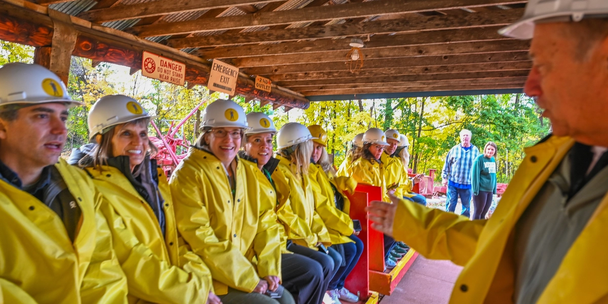 A group of people wearing bright yellow jackets and white hard hats sit on benches inside a wooden, open-sided structure with a corrugated metal roof. The setting appears to be part of a mine or industrial tour, with signs overhead reading “Danger” and “Emergency Exit.” Two people stand outside in the background near trees with autumn foliage