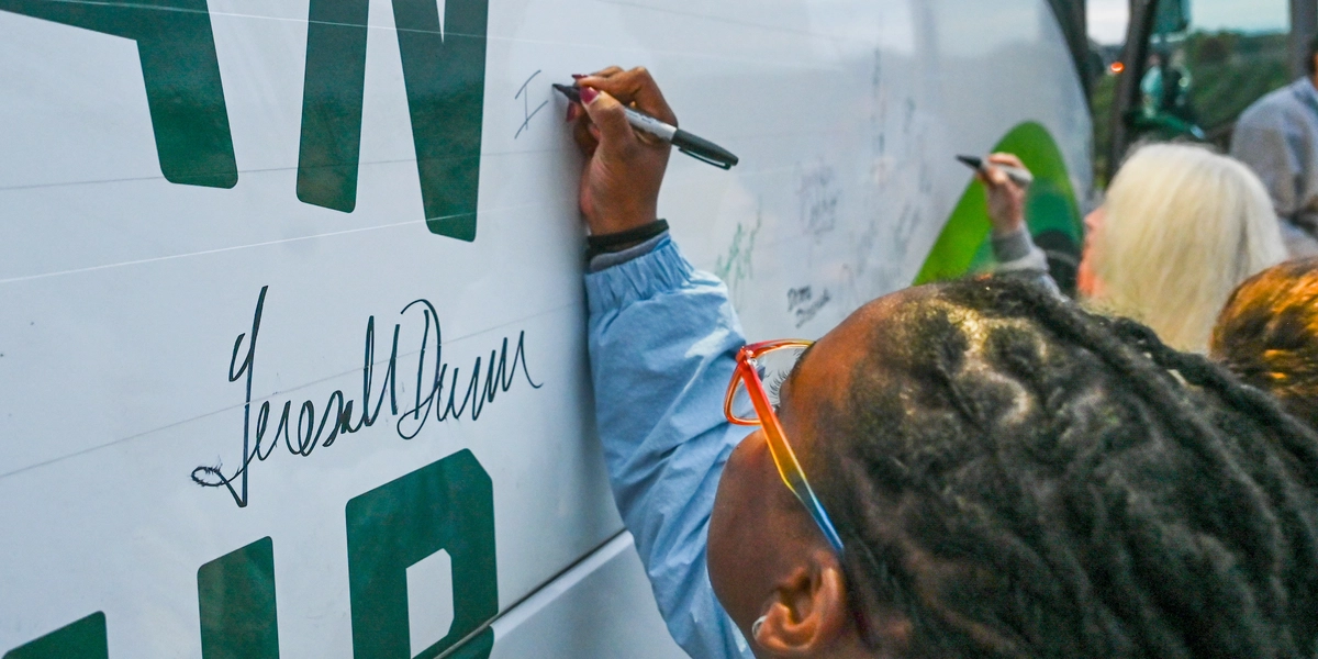 A person writes a signature on the side of a large white vehicle with green lettering using a black marker. Several other signatures are already visible on the surface. Another individual in the background is also signing the vehicle. The scene appears to be outdoors near a bus, with the activity focused on adding names or messages to the vehicle’s exterior.