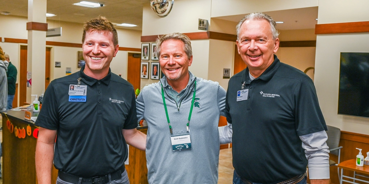 Three people stand together inside a reception area, posing for a photo. Two individuals wear black polo shirts with embroidered logos and name badges, while the person in the middle wears a gray shirt with a green lanyard and name tag. Behind them is a wooden counter decorated with orange and black Halloween-themed garland, a wall with framed photos, and a mounted TV screen to the right.
