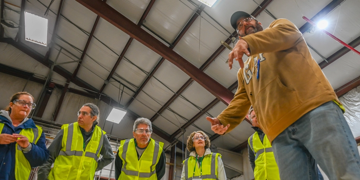 A group of people wearing bright yellow safety vests stand inside a large industrial building with a high metal ceiling and exposed beams. One person in the foreground, dressed in a tan hoodie and a black cap, gestures with one hand while speaking to the group. The background includes structural elements, fluorescent lights, and a whiteboard with notes, suggesting a tour or safety briefing in a warehouse or manufacturing facility.