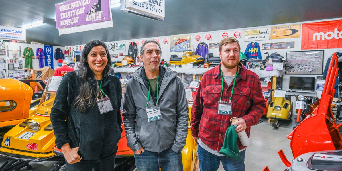 Three people stand indoors in a snowmobile museum, surrounded by vintage snowmobiles in bright colors like orange and yellow. The background features shelves and walls filled with snowmobile parts, memorabilia, and signage, including banners that read “Precision Cat,” “Boss Cat,” and “Raider Twin Snowmobile Sales & Service.” The individuals wear name badges and casual clothing, and one person holds a green cloth.