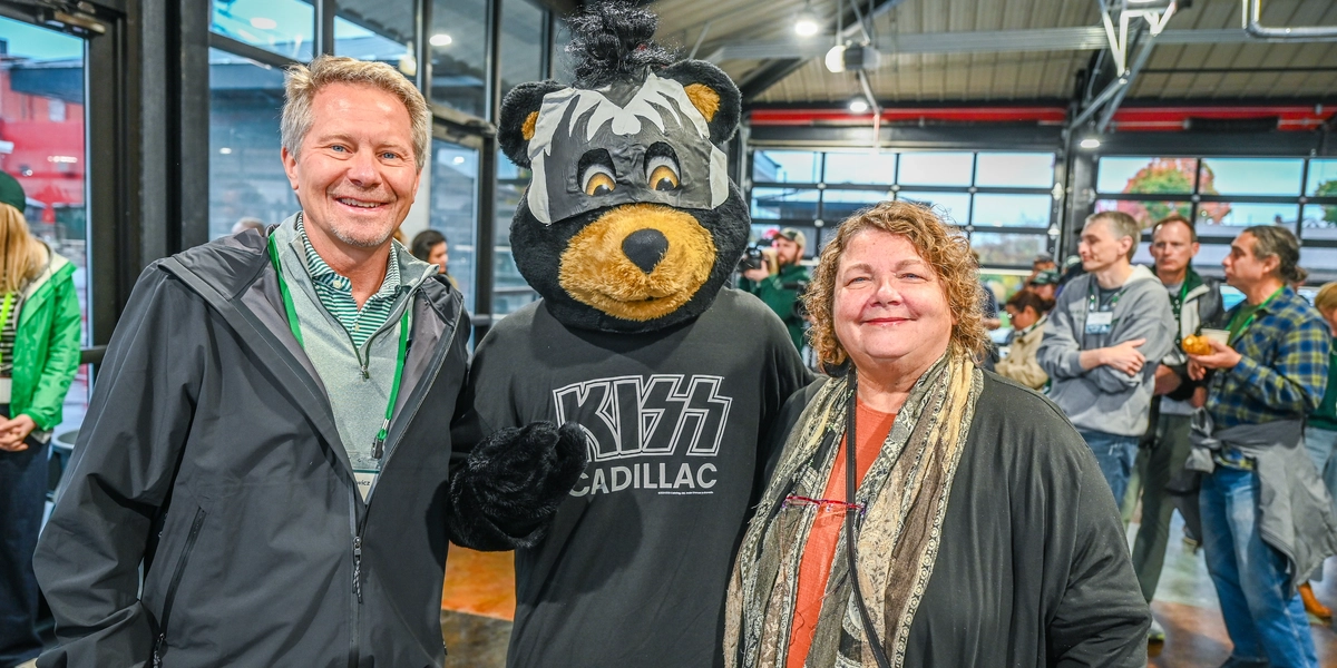 A person in a black bear mascot costume wearing a black T-shirt with the text “KISS Cadillac” stands indoors between MSU President Kevin Guskiewicz and Cadillac mayor Tiyi Schippers. The setting appears to be a large event space with high ceilings, glass garage-style doors, and a crowd of people in the background.